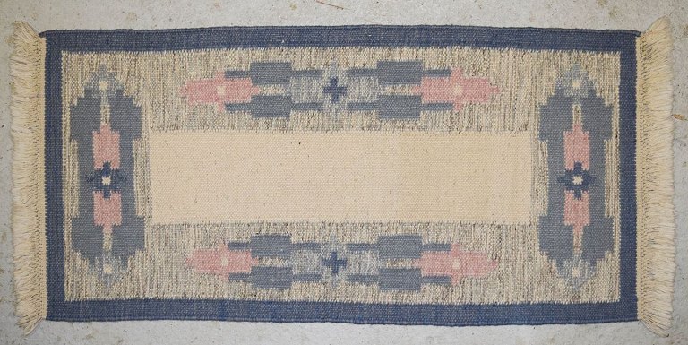 Håndvævet tæppe af uld med frynser i "Rölakan" teknik.
Geometriske former i lyseblå, mørkeblå, og lyserød på hvid baggrund.