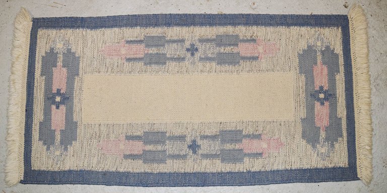 Håndvævet tæppe af uld med frynser i "Rölakan" teknik.
Geometriske former i lyseblå, mørkeblå, og lyserød på hvid baggrund.